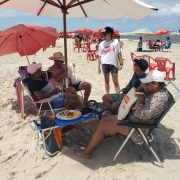 Foto horizontal colorida mostra moça de camiseta e  bone da Balada Segura em pé, conversarndo com um grupo de quatro veranistas embaixo de guarda-sóis na beira da praia