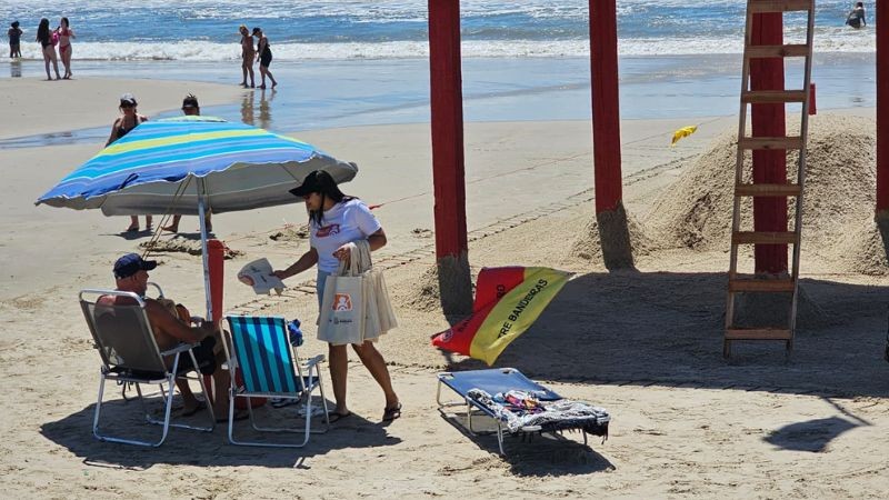 Foto horizontal colorida mostra um ambiente de praia com o mar e pessoas caminhando ao fundo. Em primeiro plano, uma mulher com a camiseta da Balada Segura entrega material edicativo para homem de boné sentado em uma cedeira de praia embaixo de um guarda sol ao lado da guarita do salva-vidas